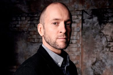 Derren Brown performing on stage, with a spotlight illuminating him and a microphone in his hand.