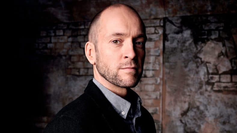 Derren Brown performing on stage, with a spotlight illuminating him and a microphone in his hand.
